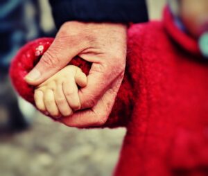 A picture of an elderly hand holding the small and of a child. The child is dressed in a red coat. The adults and child's faces can not be seen, the focus is on their hands. The picture symbolises growth, small steps, learning and learning from defeat.