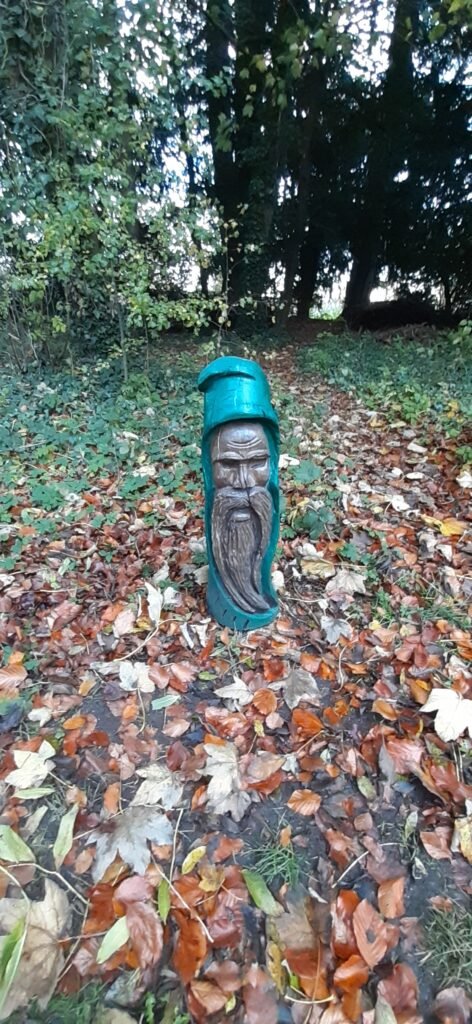 This a picture of a sculpture in Stanton park woods. The sculpture is 1 meter tall and is a depiction of a head of an old man with a long beard wearing a cyan hood. The sculpture resembles a character from the hobbit or some other folklore story. The sculpture is from the Every Cloud charity.