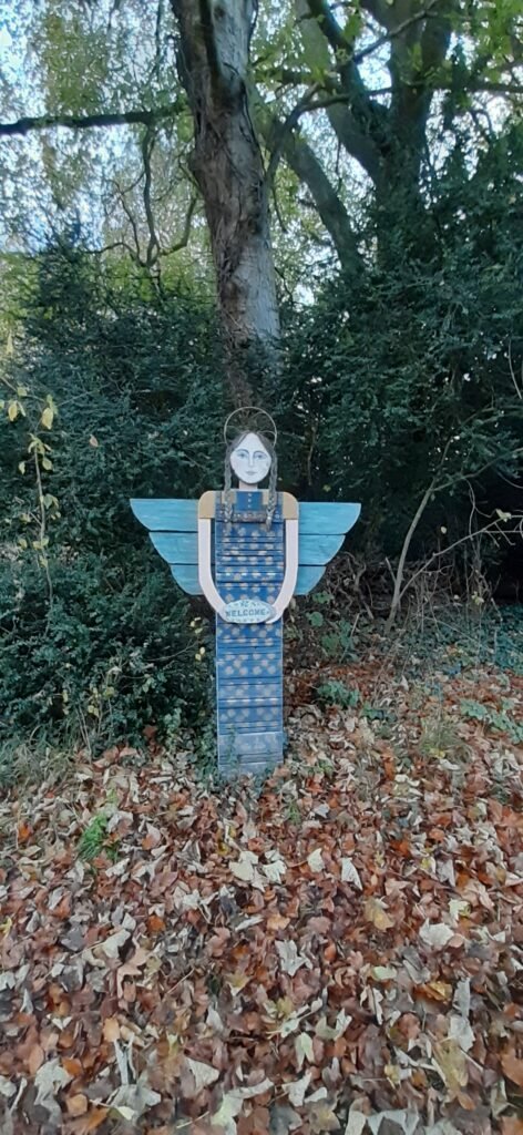 A sculpture of a girl in a blue dress with blue wings. The style of the sculptures is arts and craft with an innocence and cute feel to the sculpture. The girl looks like an angel with blue wings and is holding a welcome sign, welcoming the walkers into the woodland. The sculpture is from the Every Cloud charity.