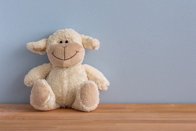 A picture of a light brown teddy bear wearing a big smile. The bear is resting against a light background and is seated in a wooden table. The picture can't help but make you smile.