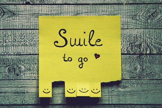 A picture of a large yellow post it note with the words "smile to go" on it in blue writing with a little heart motif. On the bottom of the post it note are little smiling faces which can be ripped off and taken. Some of these smiling faces have been already taken.