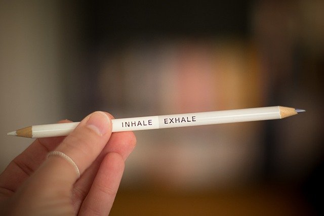 This is a picture of a hand on a fuzzy background holding a pencil. The pencil has two sharpened ends to it. On the pencil it is written "Inhale and Exhale". The aim of the picture is to promote the benefit of breath to managing stress and anxiety and to develop motivation.