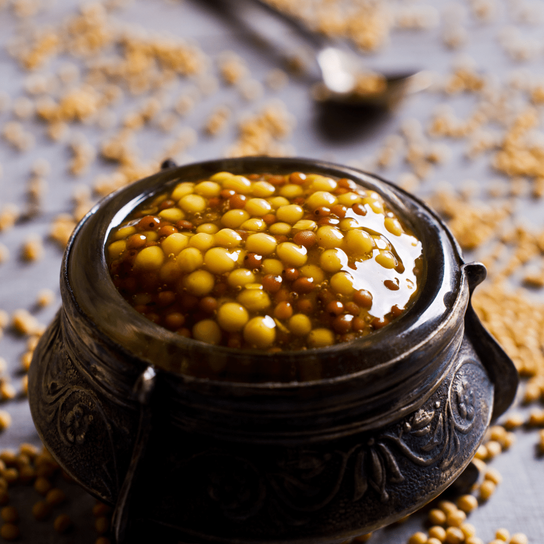 This picture has a light purple background. The main subject of the picture is a dark brown glazed pot with a wide opening. The put is the size of a small coffee cup. In the pot is yellow and brown mustard seeds in oil. Scattered around the pot are seeds and a silver spoon. The purpose of this picture is to highlight the importance of having a small amount of faith when taking action. When we have some faith that leads to taking action and small steps.