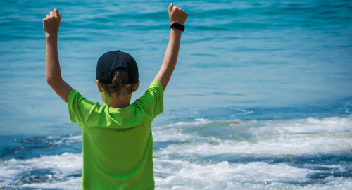 This is a picture of a boy wearing a baseball cap and a lime green T-shirt. The boy is looking out to a vivid blue sea with white surf. The boy is gesturing with his two arms up in the air showing a victory , success gesture. This is to represent confidence and how Hypnotherapy Swindon can help.