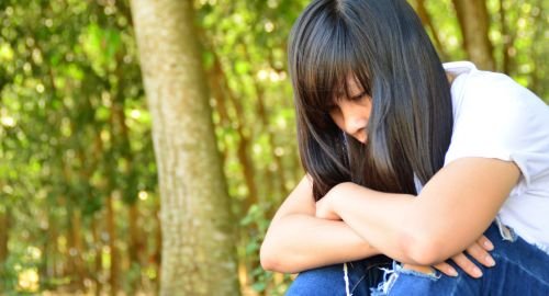 This is a picture with a blurred woodland background of a woman, wearing blue jeans and a white T-shirt. The woman is sat down, her arms on her knees looking depressed. The purpose of the picture is to convey depression and that Hypnotherapy Swindon can help with depression.