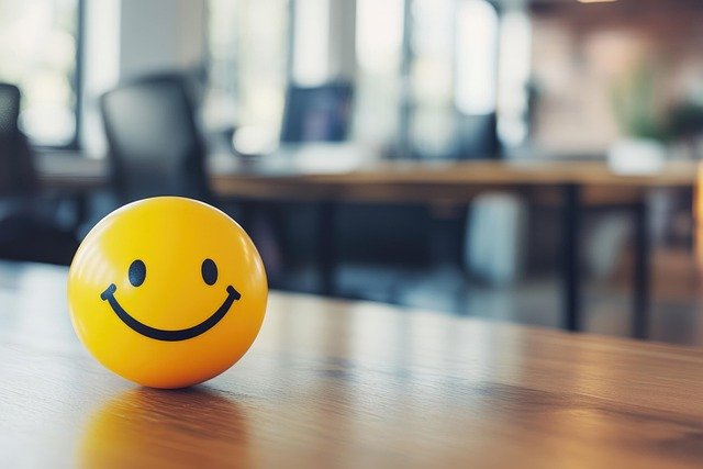 A blurred office background. In the foreground is a table upon which sits a yellow smiley ball. This represents the benefits of mindfulness, it makes you happy.